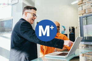 logo van M+ op een foto van twee mannen die naar een laptop kijken, waarvan er 1 aan het wijzen en uitleggen is